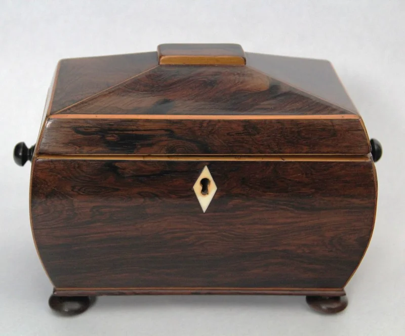 Theekistje in palissanderhout - Diversen › Dozen en theekistjes - Sarcofaagvormig theedoosje in palissanderhout en afboordingen in satijnhout. Binnenin voorzien van twee compartimenten, elk afgedekt met een dekseltje in palissanderhout en een knopje in ivoor.
22 x 13cm, 16cm hoog
Georgean periode, ca 1820
Gepolitoerd op oude wijze