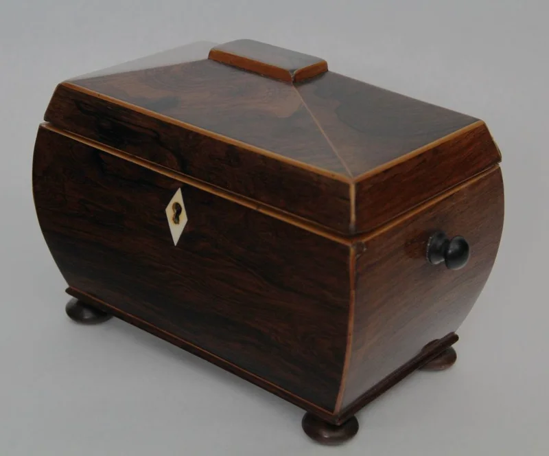 Theekistje in palissanderhout - Diversen › Dozen en theekistjes - Sarcofaagvormig theedoosje in palissanderhout en afboordingen in satijnhout. Binnenin voorzien van twee compartimenten, elk afgedekt met een dekseltje in palissanderhout en een knopje in ivoor.
22 x 13cm, 16cm hoog
Georgean periode, ca 1820
Gepolitoerd op oude wijze